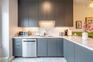 Een moderne keuken met grijze kastjes, een roestvrijstalen vaatwasser, een magnetron, een koffiezetapparaat en een bloevas op het aanrecht.