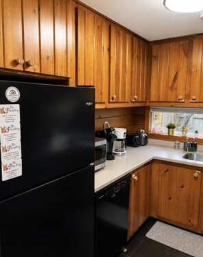 A kitchen with wooden cabinets, a black refrigerator, a coffee maker, and a window with plants. - Sea La Vie - Hot Tub, Huge Enclosed Private Deck, Sleeps 22, Close To Town (Ocean Beach)