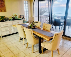A dining area with a wooden table, four chairs, and a view of the ocean through large windows. - Oceanfront Angel Villa with dipping pool, dog friendly and secure garage (B.C.)