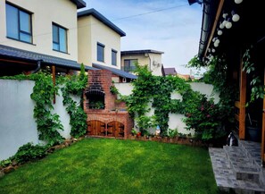 A residential backyard with a brick fireplace, a grassy lawn, and a white wall with climbing plants. - A cosy villa with private garden and free parking, on the outskirts of Bucharest (Bragadiru)