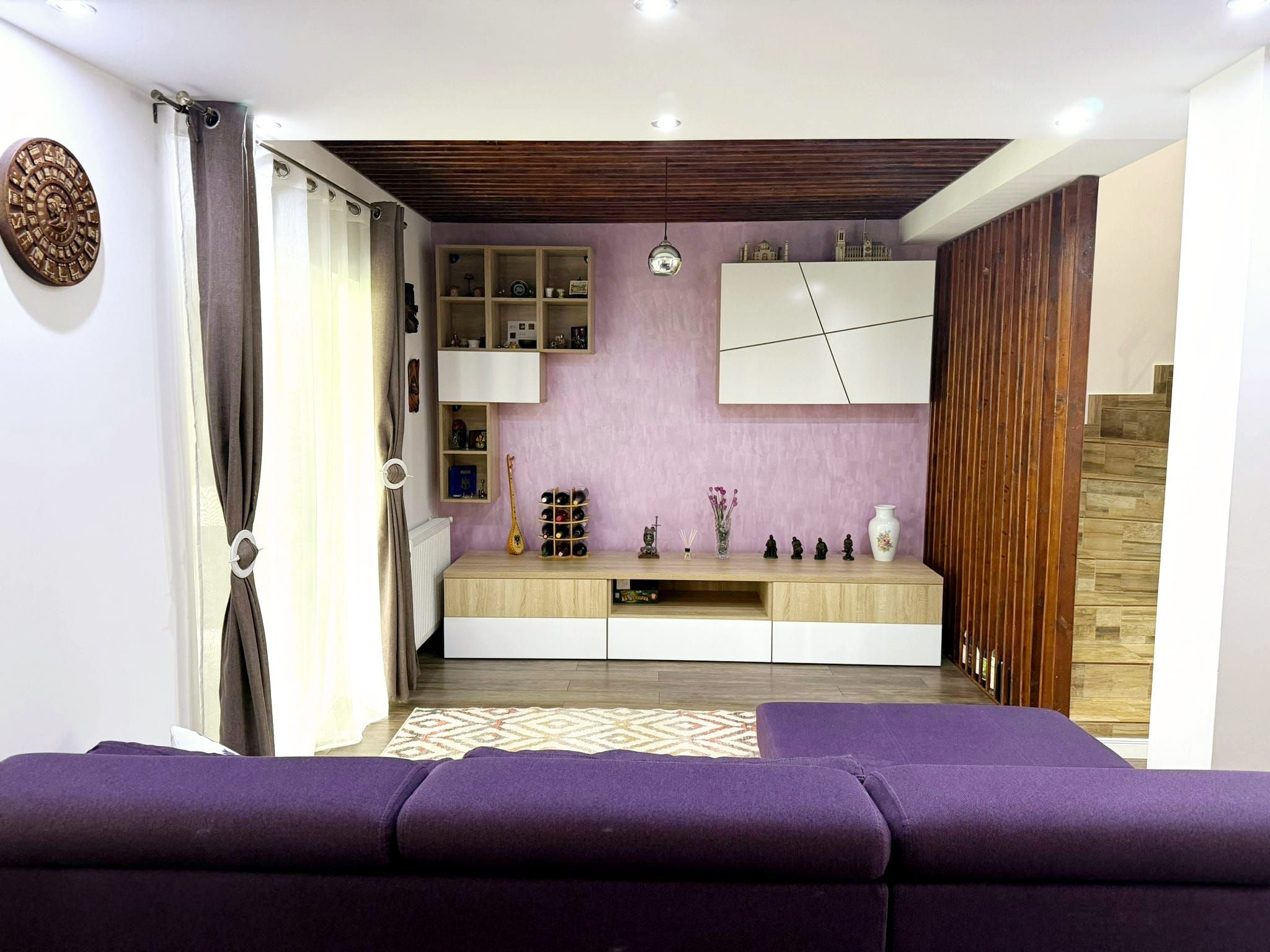 Ein modernes Wohnzimmer mit einem violetten Sofa, einer Holz-Unterhaltungsecke und einer Küchenzeile mit Schränken und einem Weinkühlschrank.