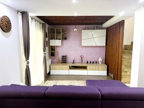 A modern living room with a purple sofa, a wooden entertainment unit, and a kitchen area with cabinets and a wine rack.