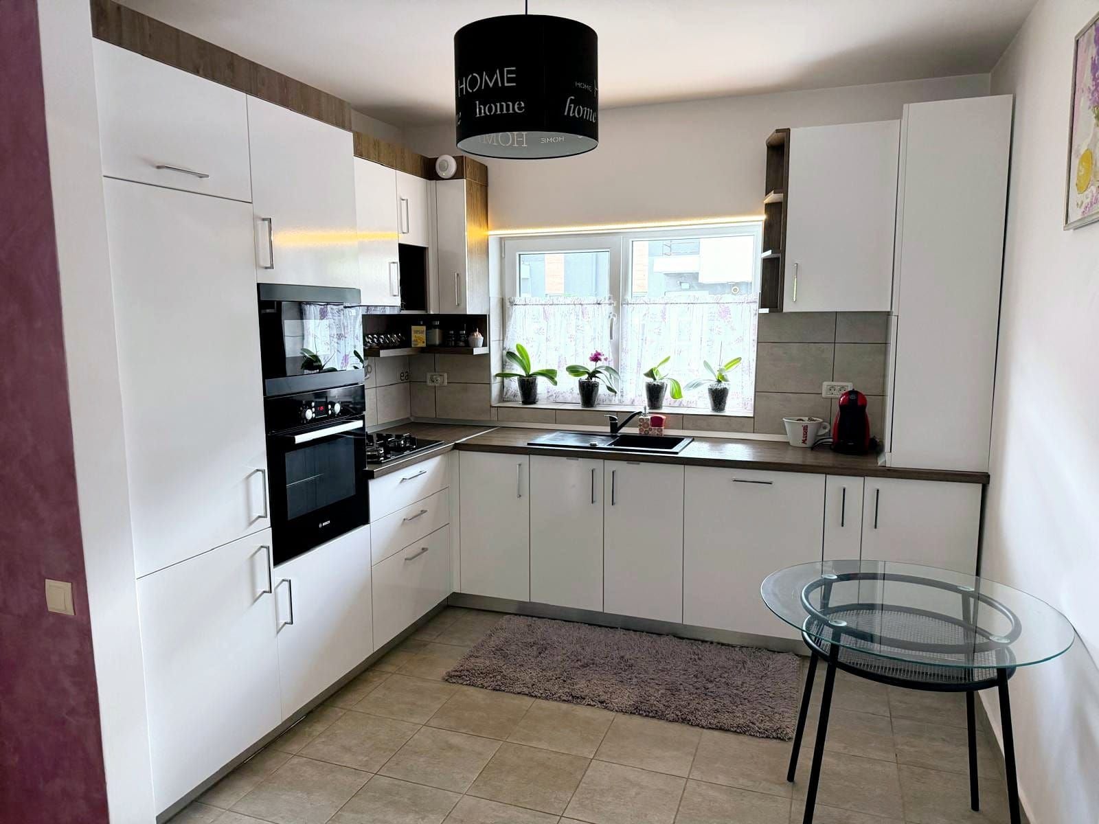 Una cocina moderna con armarios blancos, una estufa negra, una mesa de vidrio y una ventana con plantas.