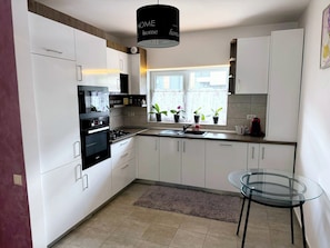 A modern kitchen with white cabinets, a black stove, a glass table, and a window with plants.