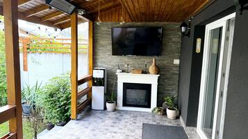 Un patio cubierto con un televisor de pantalla plana montado en la pared, una pequeña chimenea blanca y un rincón de estar con un banco y una mesa.