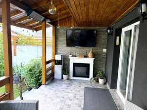 A covered patio area with a flat-screen TV mounted on the wall, a small white fireplace, and a seating area with a bench and a table. - A cosy villa with private garden and free parking, on the outskirts of Bucharest (Bragadiru)
