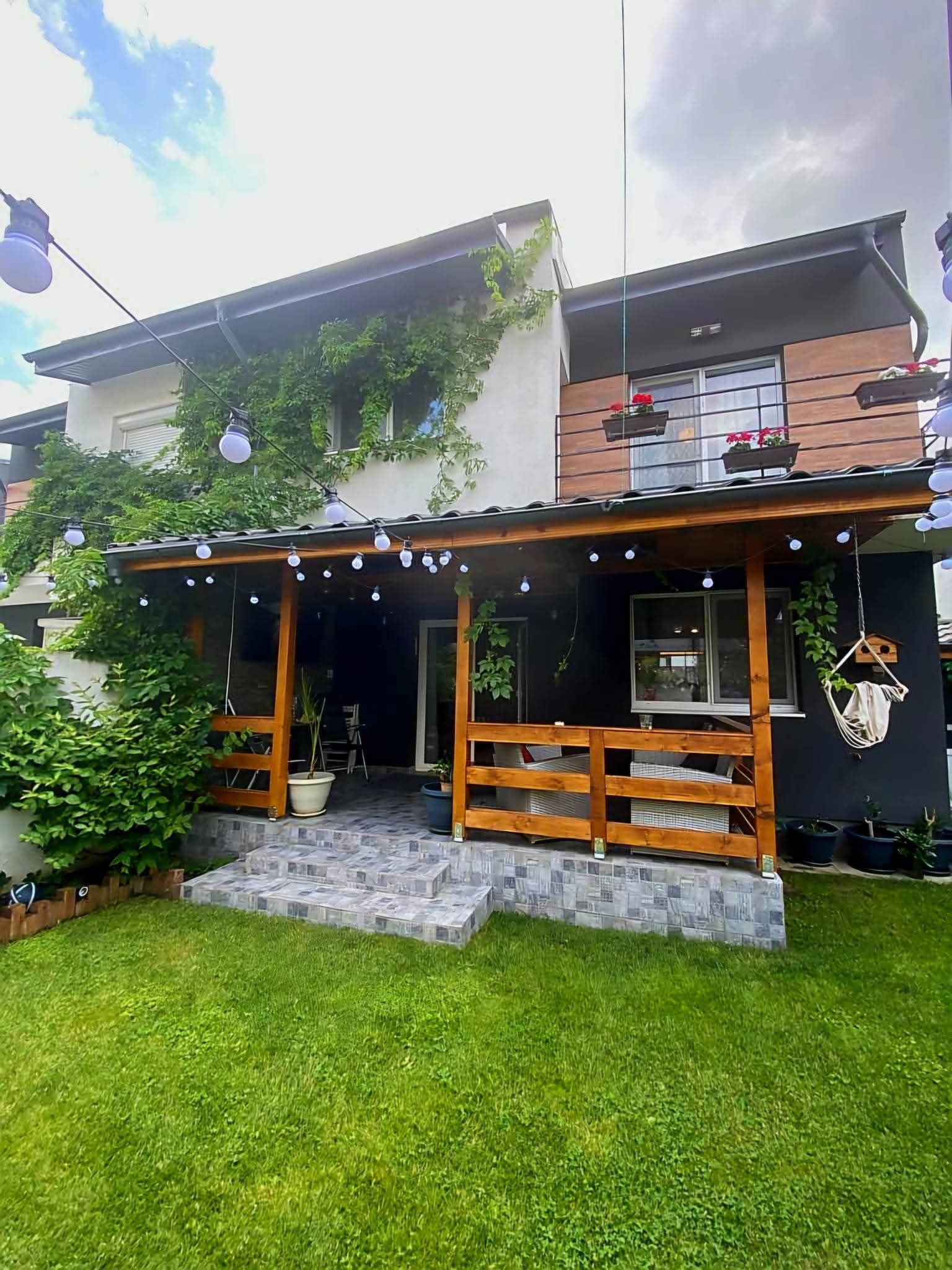 Una casa de dos pisos con un porche de madera, plantas en macetas y luces colgantes.