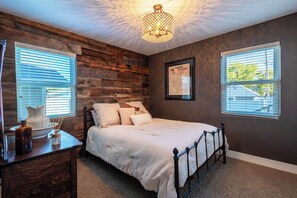A bedroom with a wooden headboard, a bed with white bedding, a nightstand with a lamp, and a window with blinds. - 4-bedroom house with pool and hot tub.  Walking distance to downtown Spearfish. (Spearfish)
