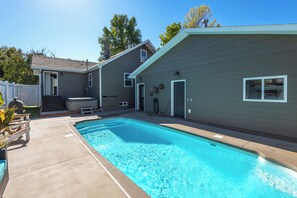 Une piscine résidentielle avec un jacuzzi, un patio couvert et une maison à l’extérieur grise.