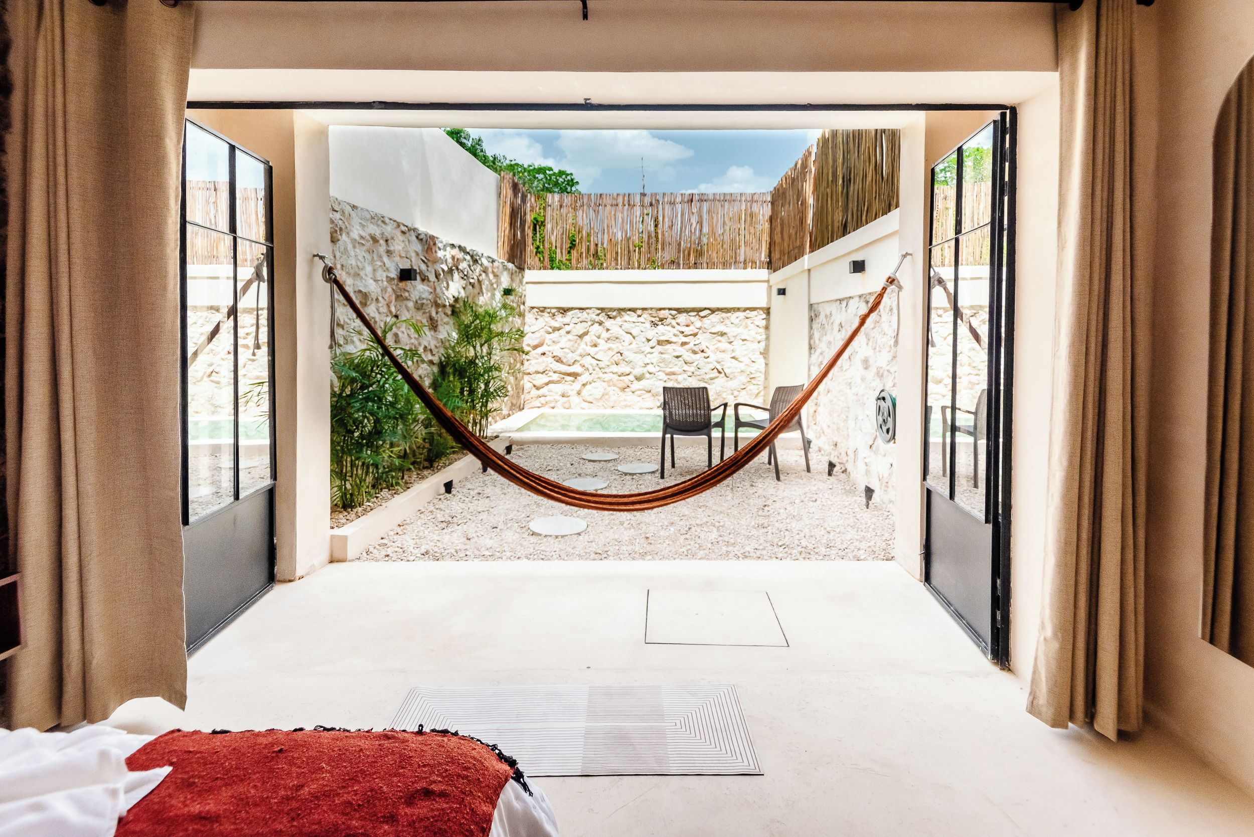 A room with a hammock, a bed, and a view of an outdoor patio with a stone wall and plants.