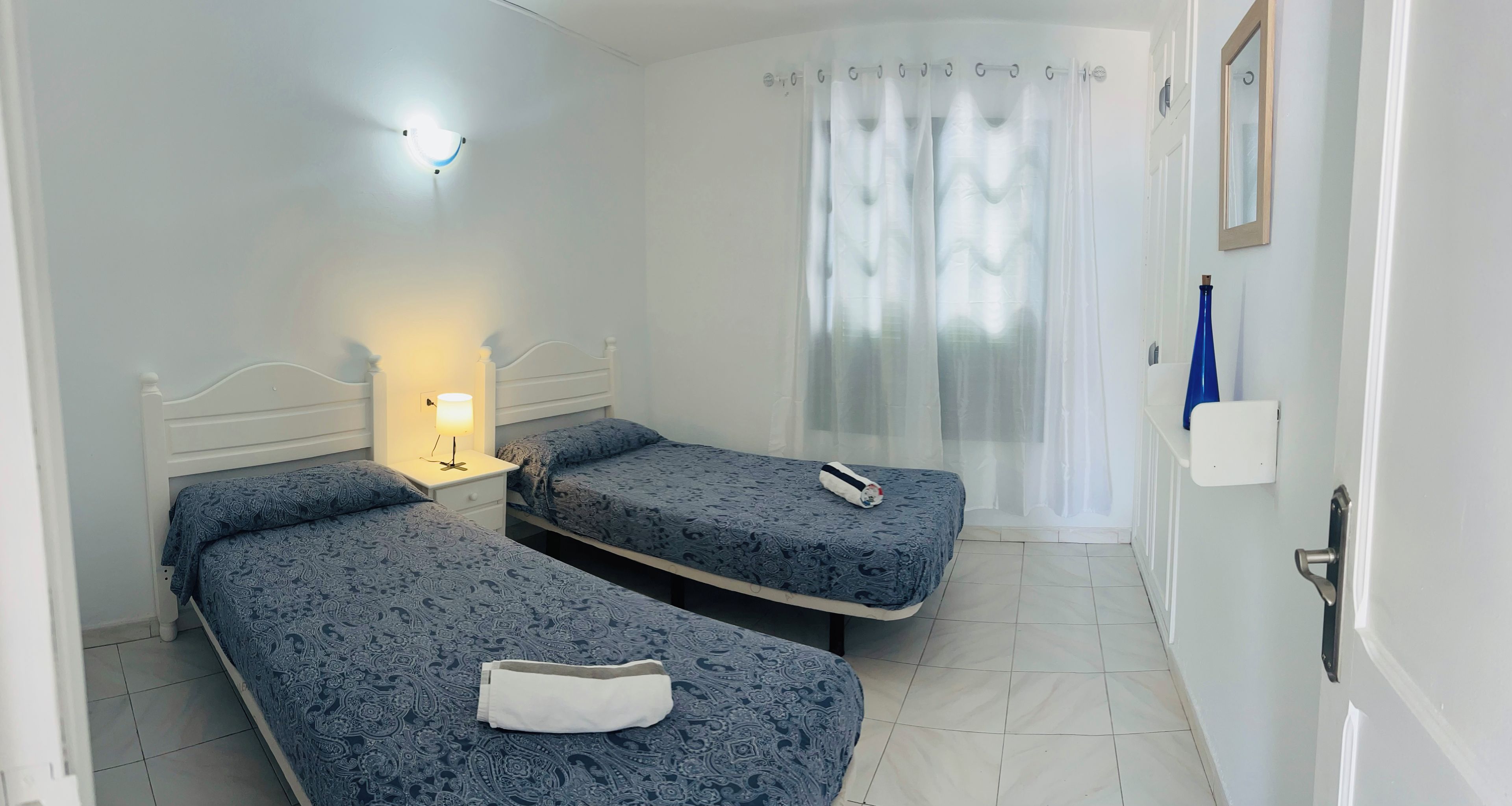 Habitación de hotel con dos camas individuales, una mesita de noche con lámpara, un baño con cortina de ducha y una puerta con manija.