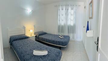 Habitación de hotel con dos camas individuales, una mesita de noche con lámpara, un baño con cortina de ducha y una puerta con manija.