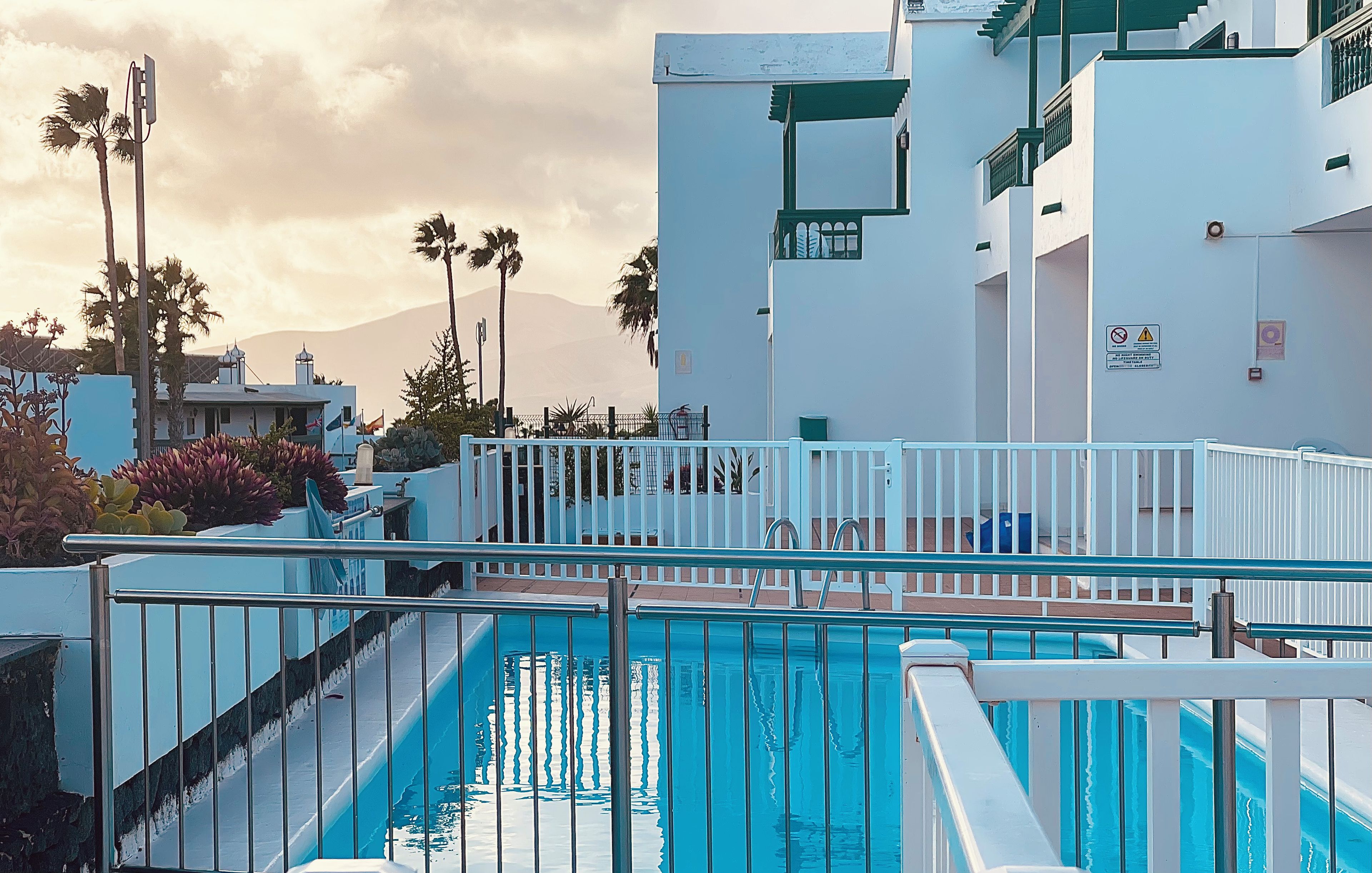 Una piscina con baranda metálica, rodeada de edificios con paredes blancas y detalles verdes, y palmeras al fondo.