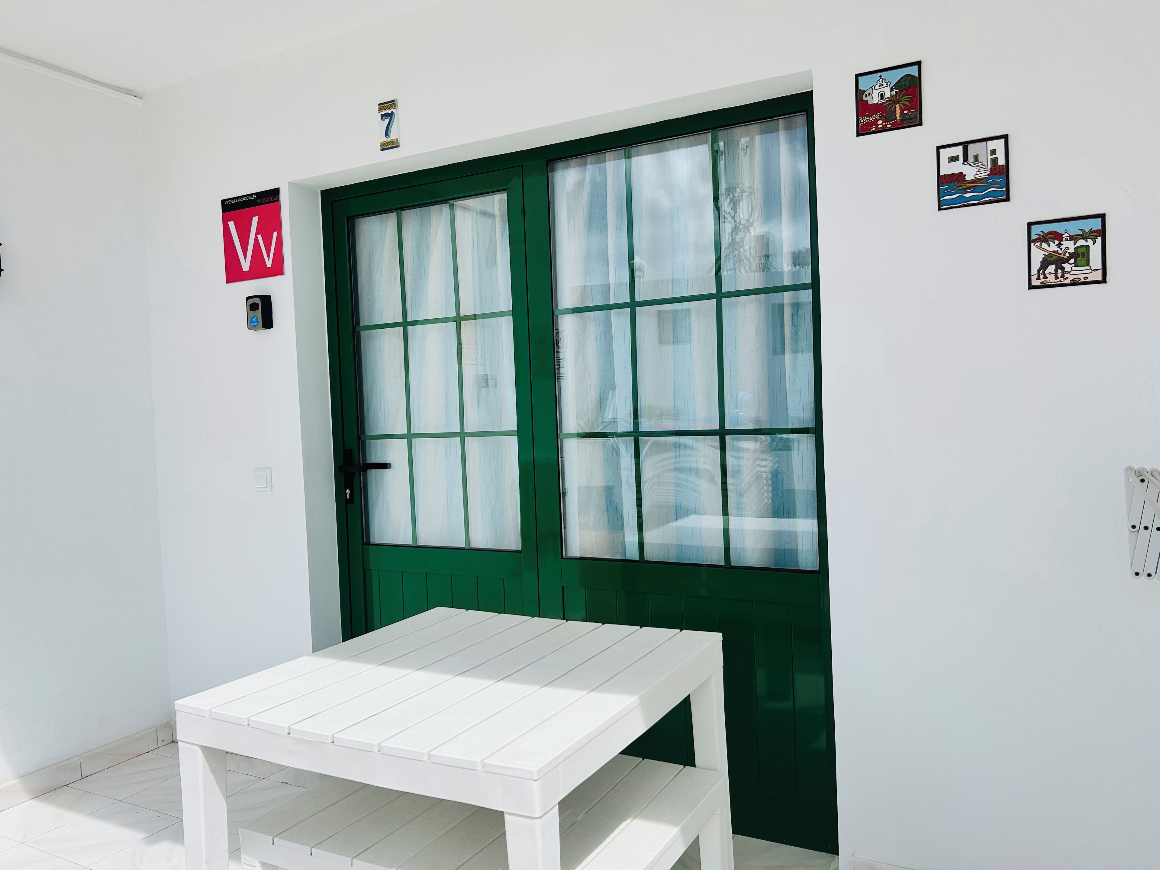 Een kamer met een groene deur, een witte tafel en een witte muur met enkele afbeeldingen.