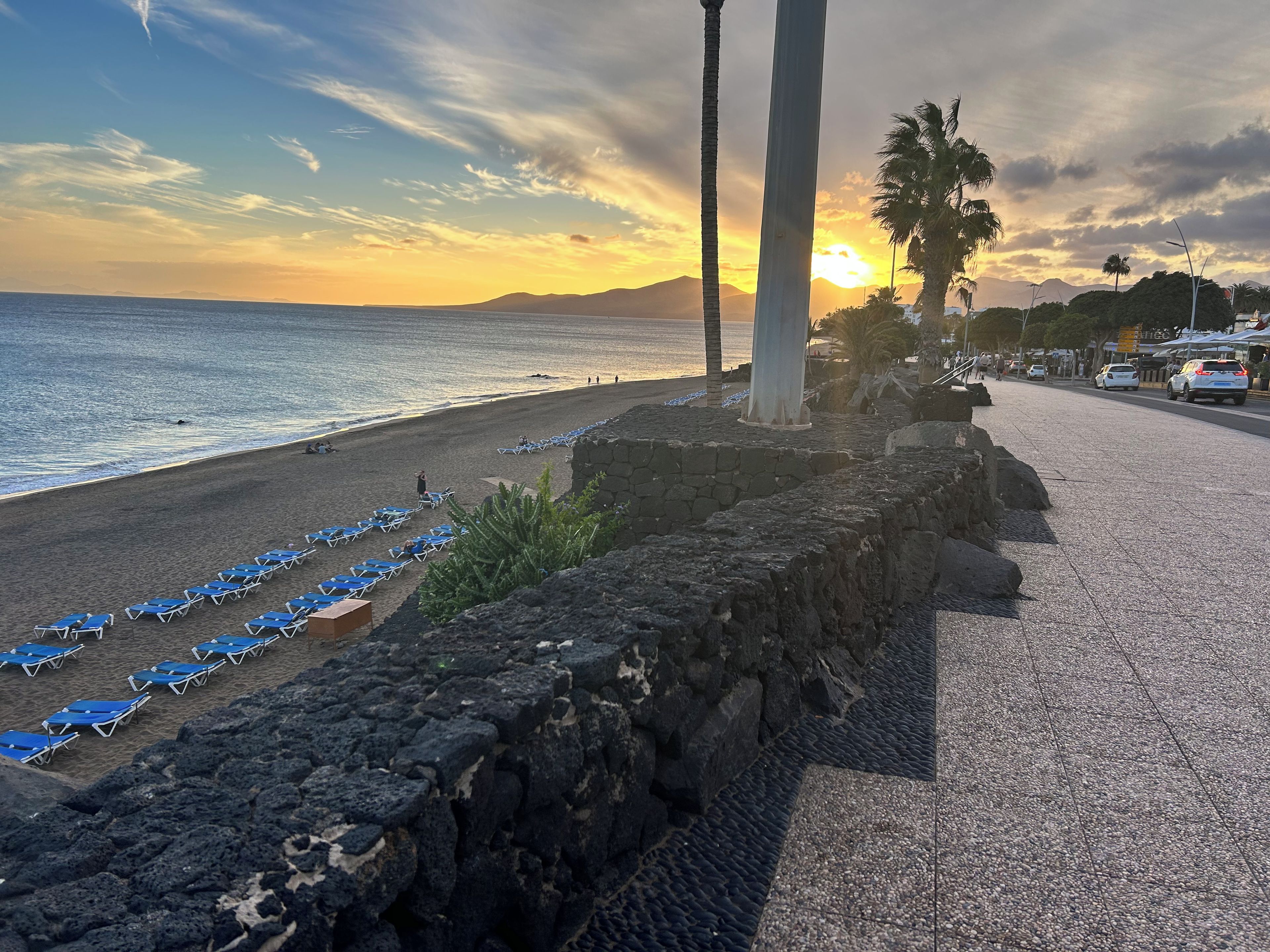 Una playa con hileras de sillas azules, un muro de piedra y una vista despejada del océano y el atardecer.