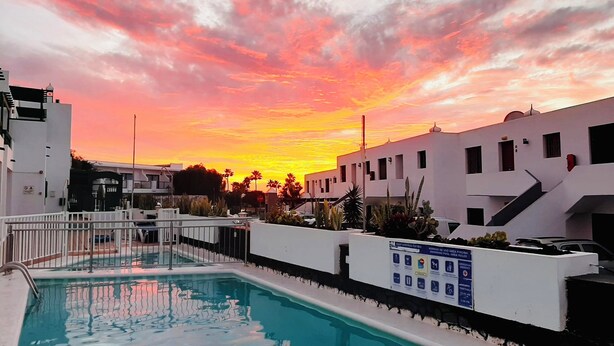 En swimmingpool med udsigt til solnedgang, omgivet af hvide bygninger og palmer.