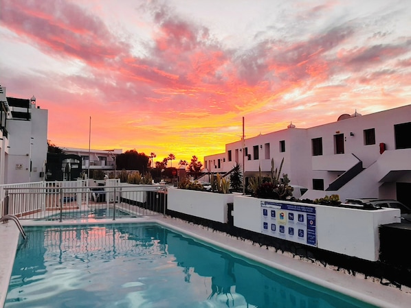 Ein Swimmingpool mit Blick auf einen Sonnenuntergang, umgeben von weißen Gebäuden und Palmen.