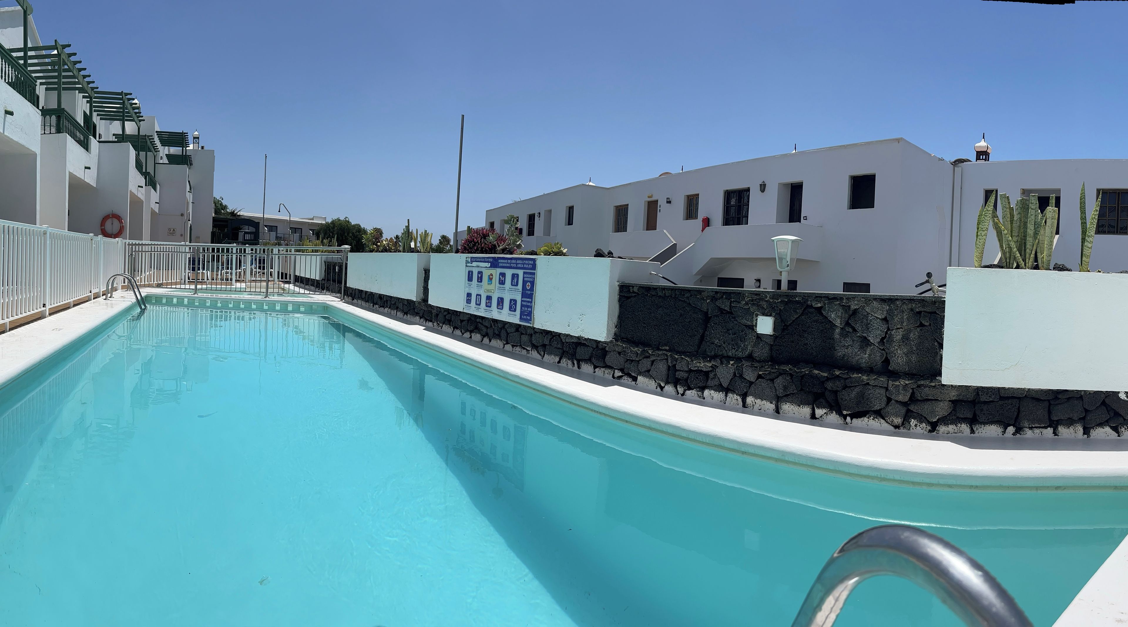 Une piscine bordée d’un mur en pierre et d’une rambarde en métal, entourée de bâtiments blancs, avec un ciel dégagé au-dessus.