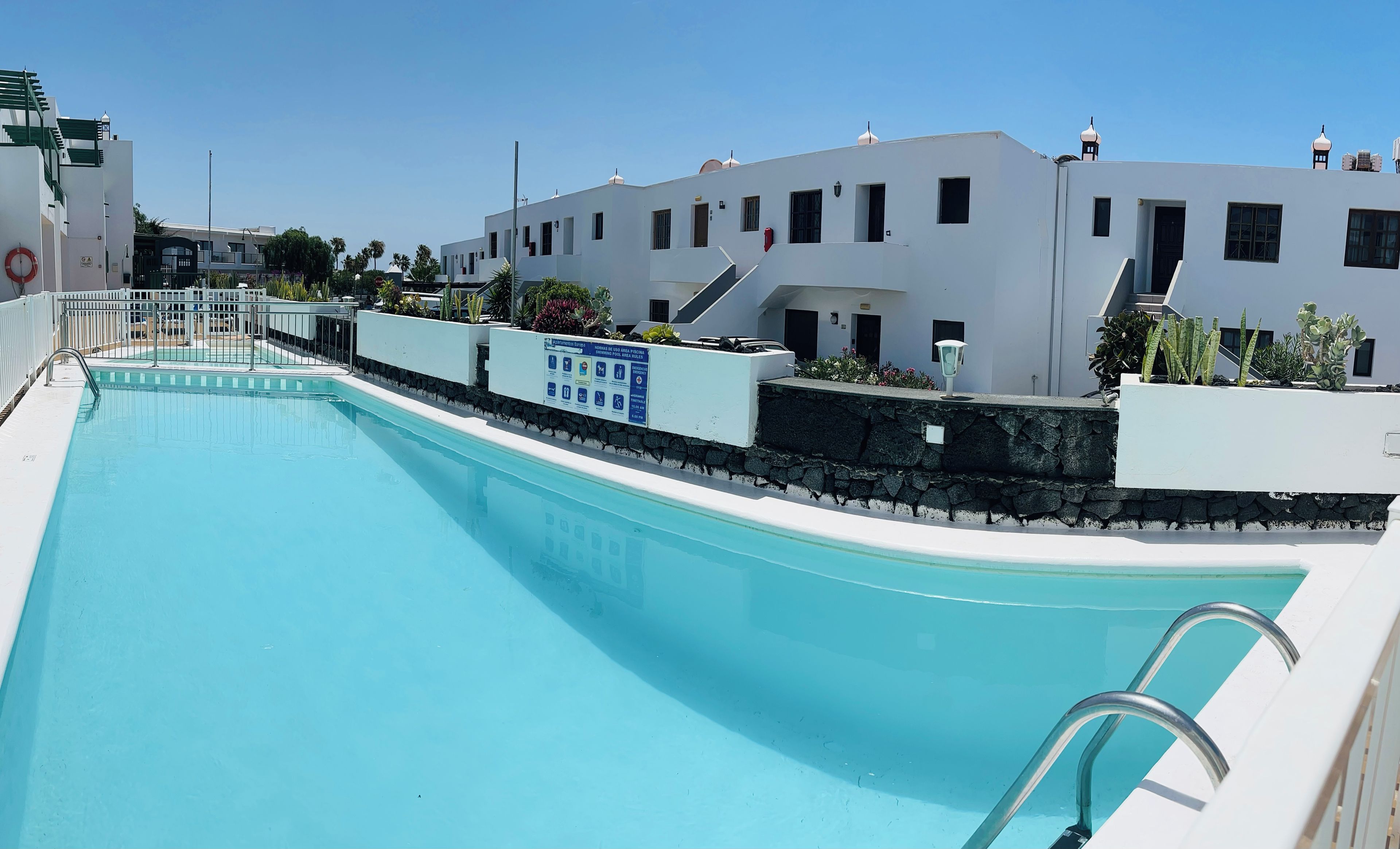 Una piscina con un muro de piedra y barandillas metálicas, rodeada de edificios blancos con numerosas ventanas y balcones.