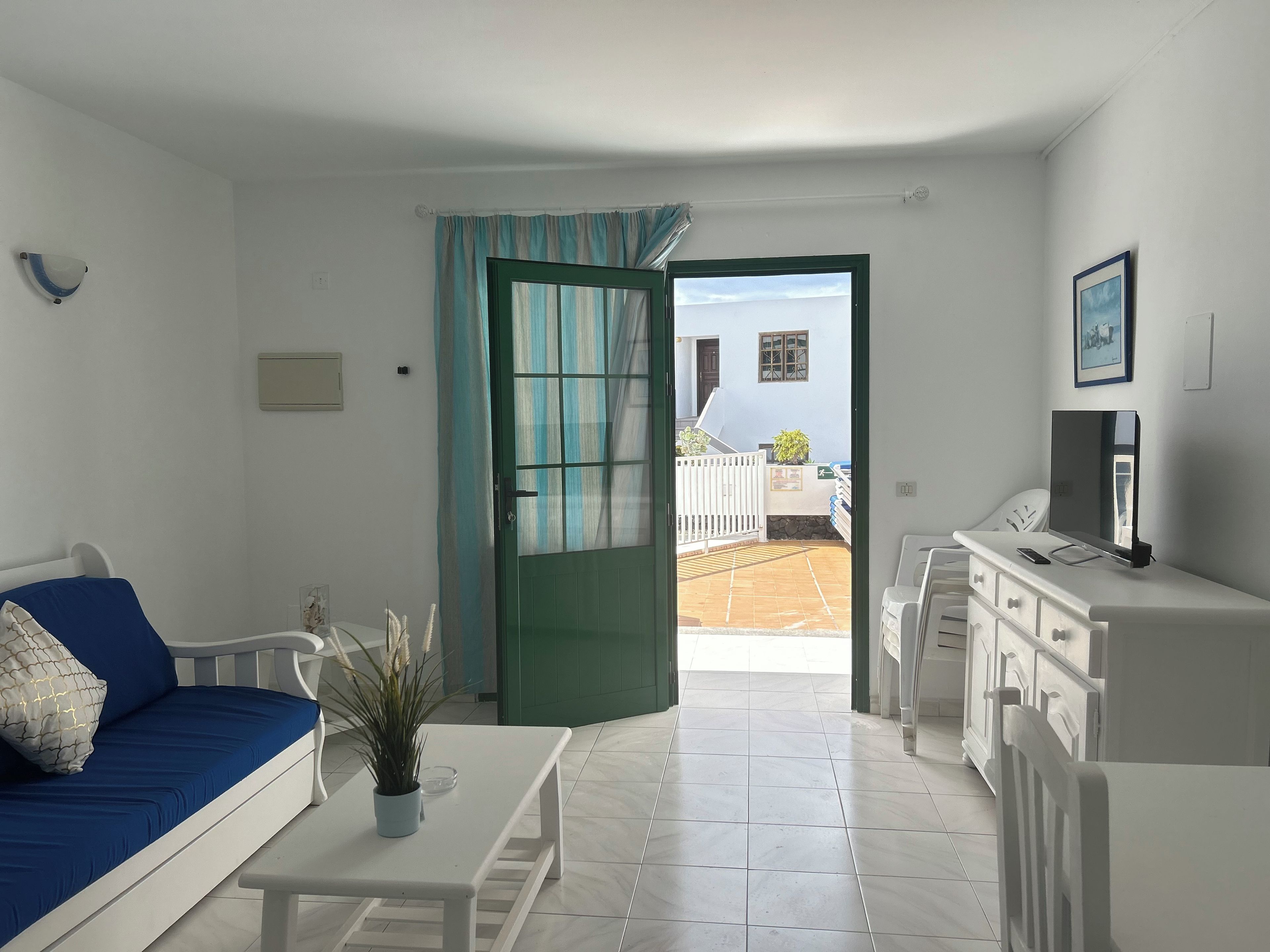 Una habitación moderna y bien iluminada con un sofá blanco, una mesita con una planta, un televisor sobre un mueble y vistas a un patio a través de una puerta abierta.