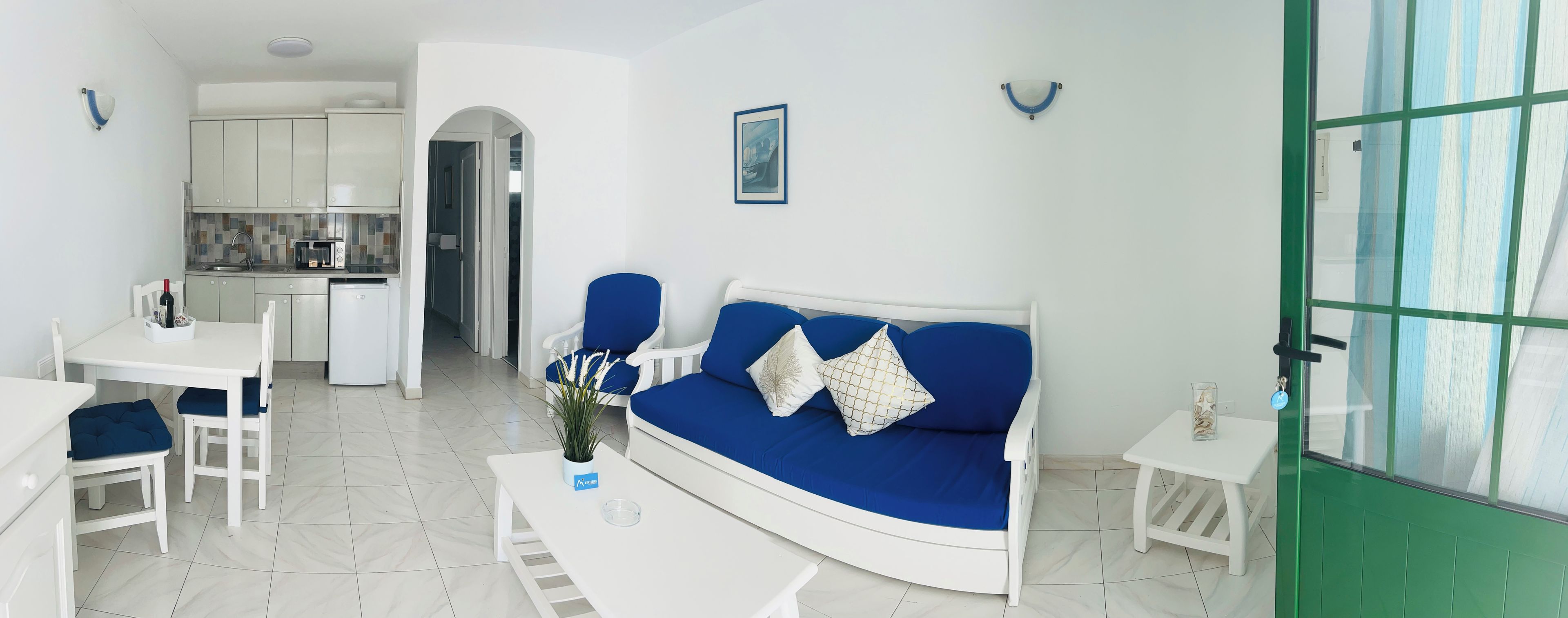 Un espacio habitable compacto con kitchenette, un sofá con cojines azules, una mesa de comedor con sillas y una pequeña mesita auxiliar.