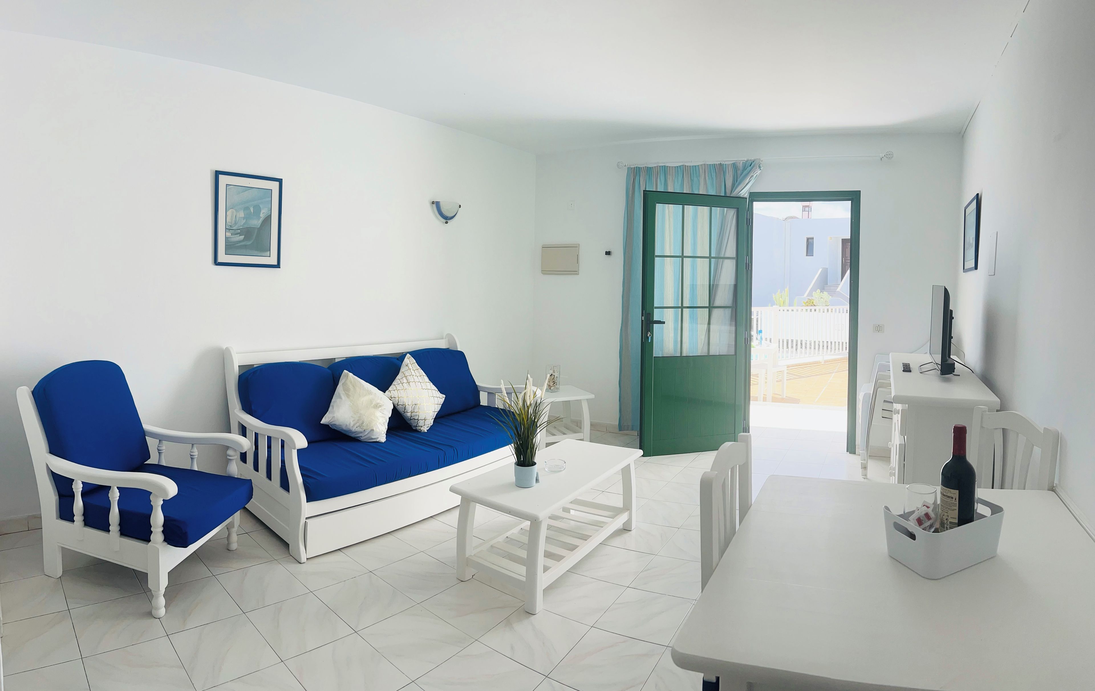 Una sala de estar con un sofá blanco, una mesa de centro blanca y una mesa de comedor. Hay una puerta verde que da a un espacio exterior.