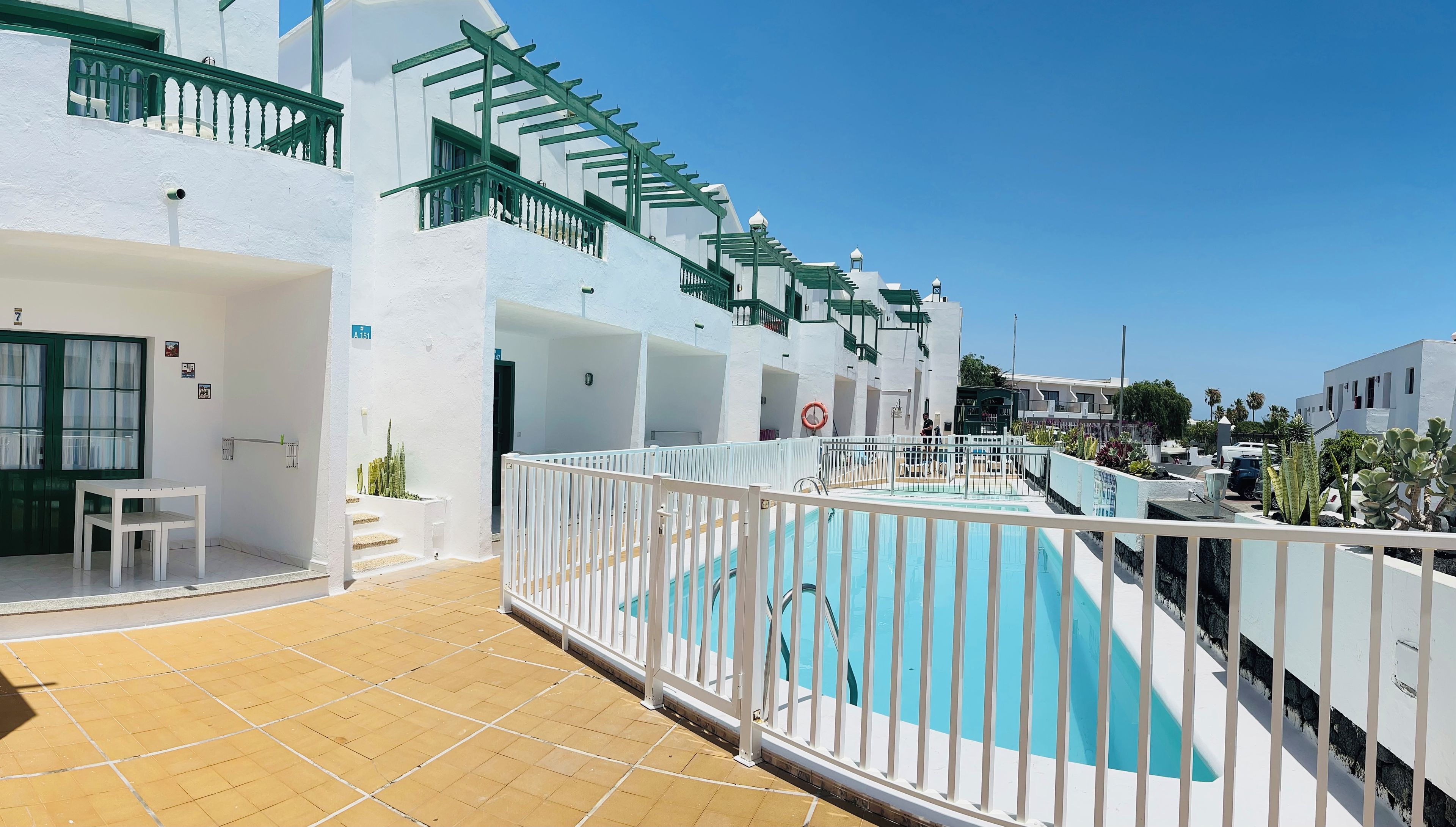 Un espace piscine entouré d’une clôture blanche, avec un escalier et un bâtiment aux murs blancs et aux balustrades vertes.