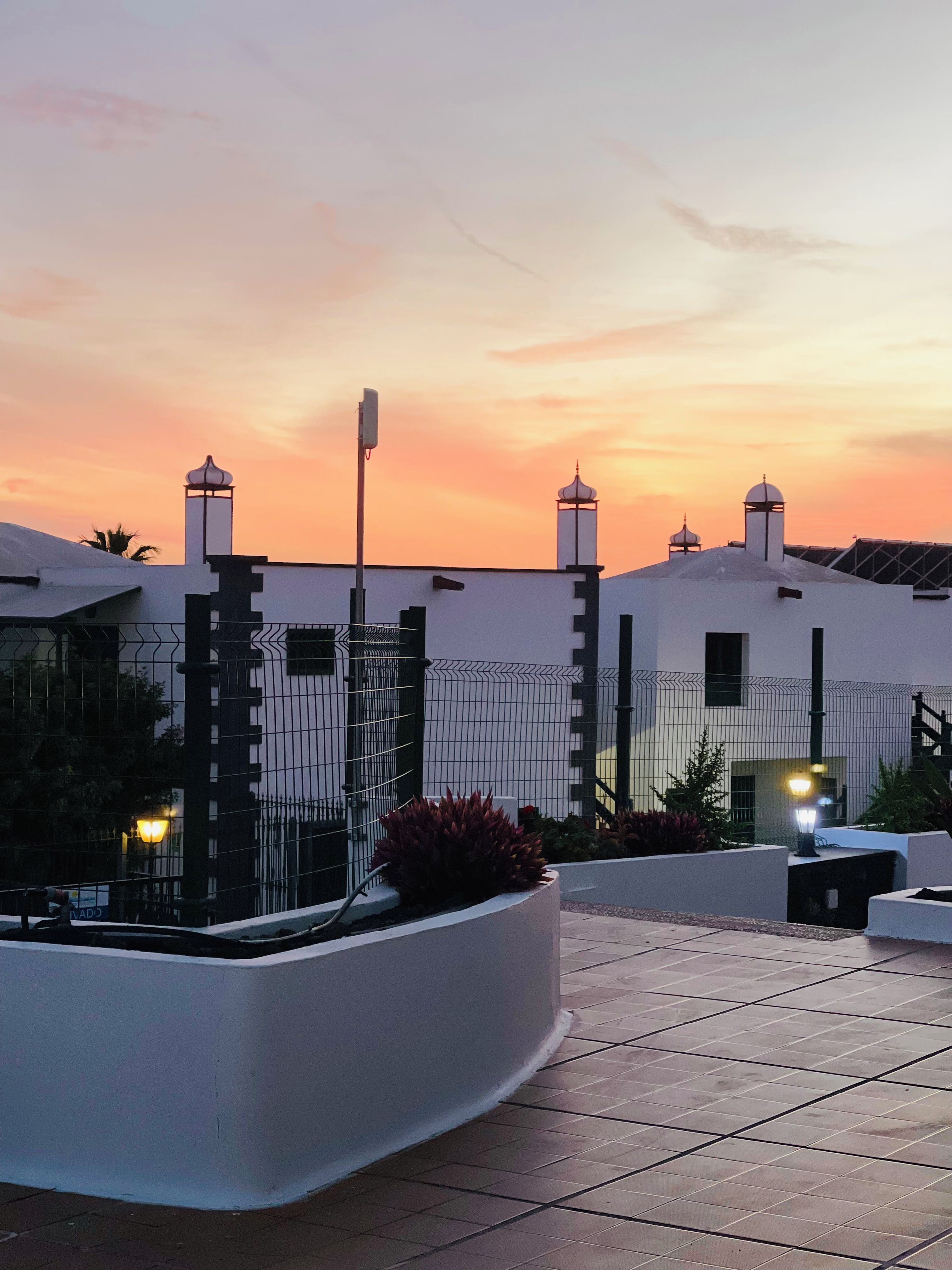 A rooftop terrace with a white building, a fence, and a sunset sky.