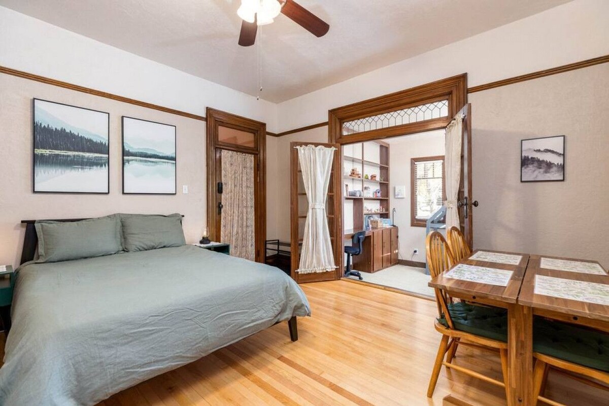 Ein Schlafzimmer mit einem Bett, einem Schreibtisch und einem Sessel. Es gibt zwei Fenster und eine Tür zu einem anderen Raum.