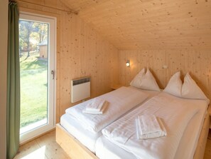 A double bed with white linens and pillows, a window with curtains, and a wall-mounted heater. - One-bedroom Cabin (Sankt Georgen am Kreischberg)