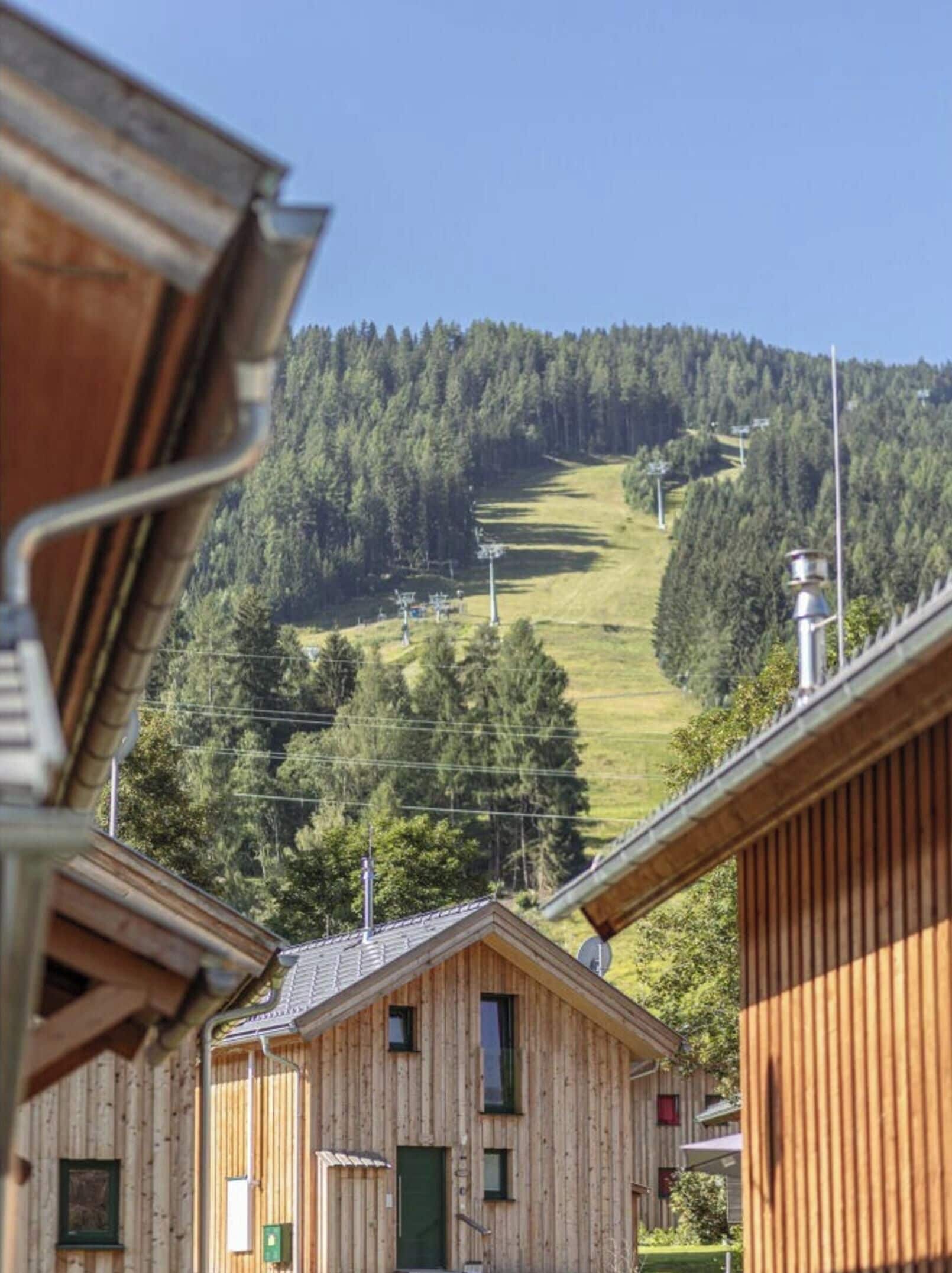 A wooden cabin with a view of a forested hill and a ski lift in the background.