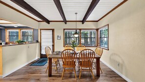A dining room with a wooden table and chairs, large windows, and a kitchen area with a sink and countertop. - Pines & Peace by Avantstay Log Cabin + Wraparound Deck + Grill Mins to Ski & Lake (Truckee)
