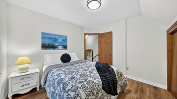 Quarto com cama, mesa de cabeceira, luminária, pintura e porta de madeira.