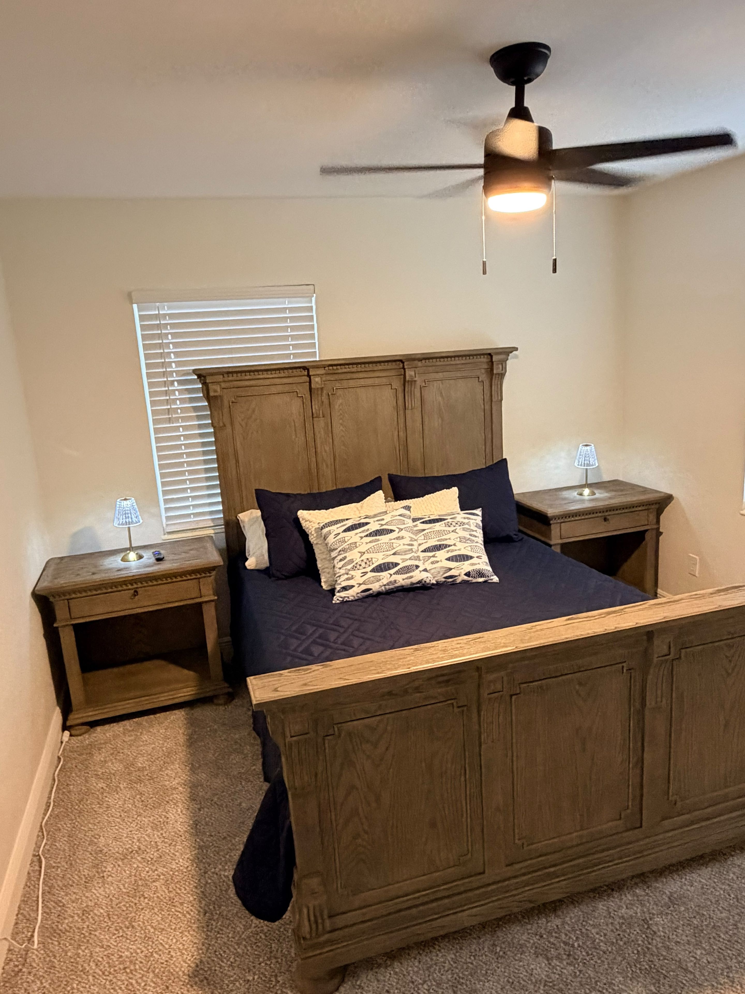 A bedroom with a wooden bed, two bedside tables, a ceiling fan, and a window with blinds.