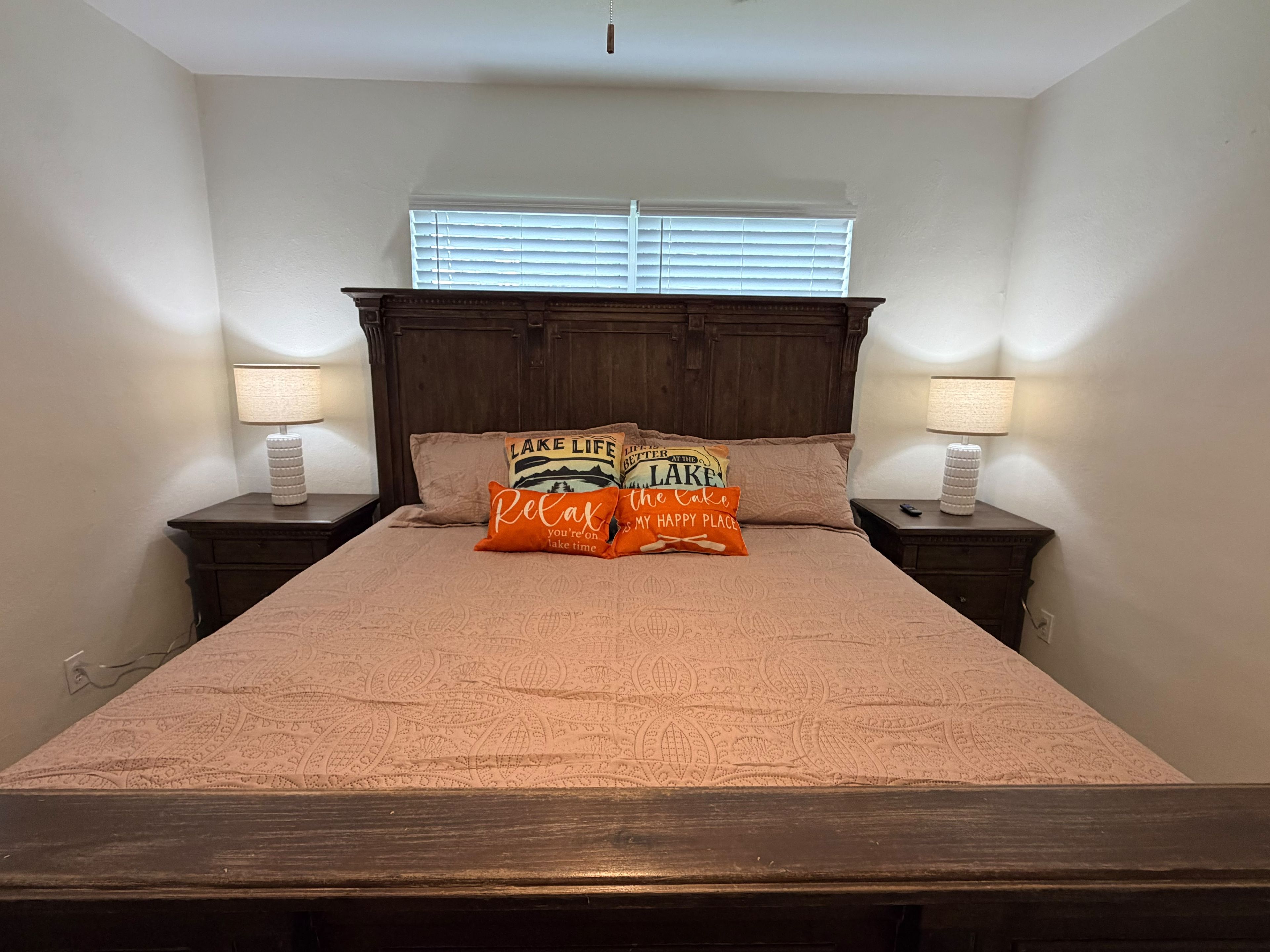 A bedroom with a wooden headboard, two bedside lamps, a bed with patterned bedding, and pillows with text.