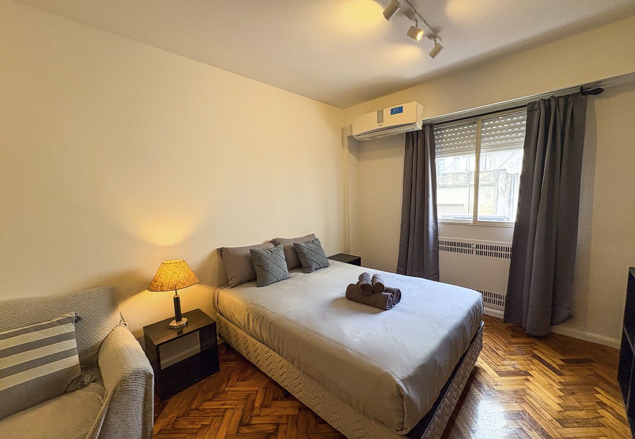 A hotel room with a bed, a nightstand with a lamp, a window with curtains, and a radiator.