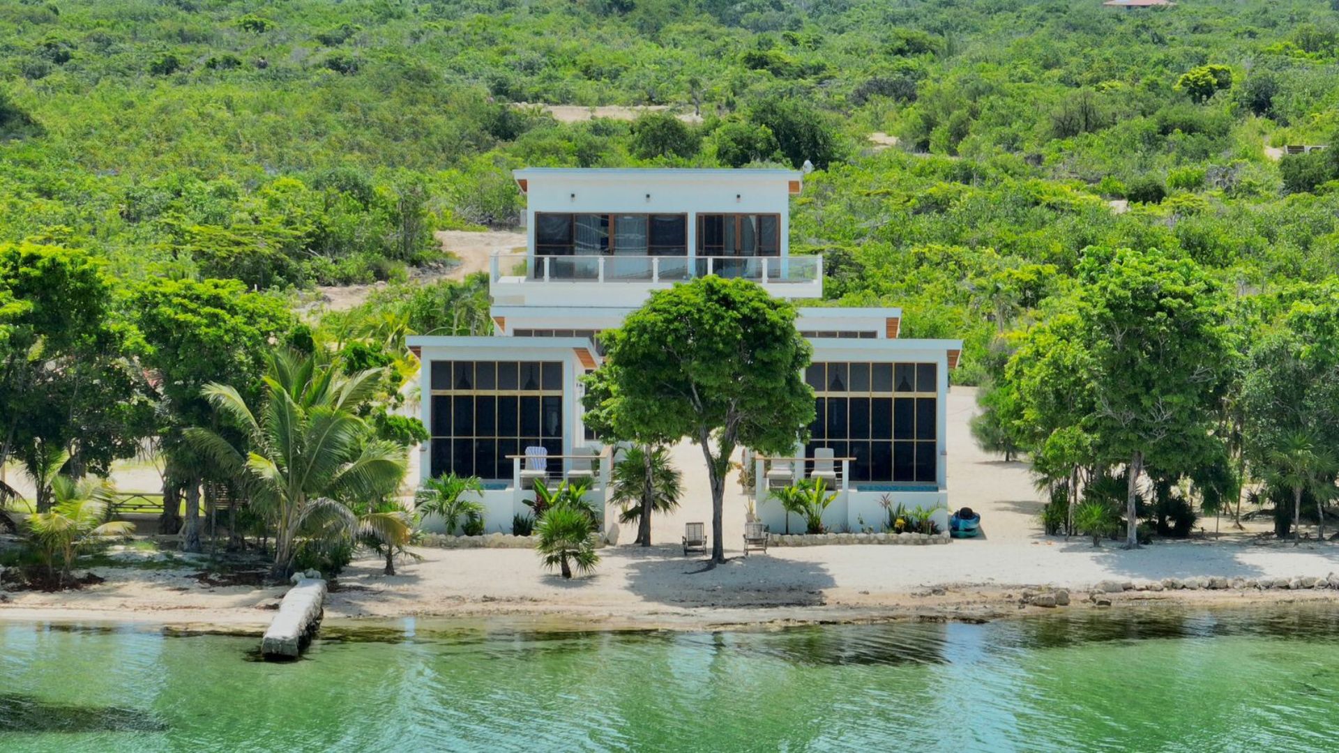 A modern two-story house with a large balcony, surrounded by lush greenery and a body of water.
