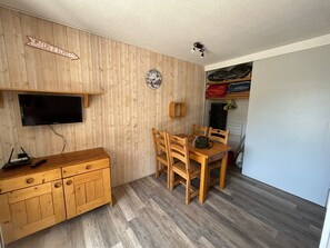 A compact room with a wooden wall, a mounted television, a dining table with chairs, and a coat rack with folded clothes. - 1800 Village Studio with a View (Les Deux Alpes)
