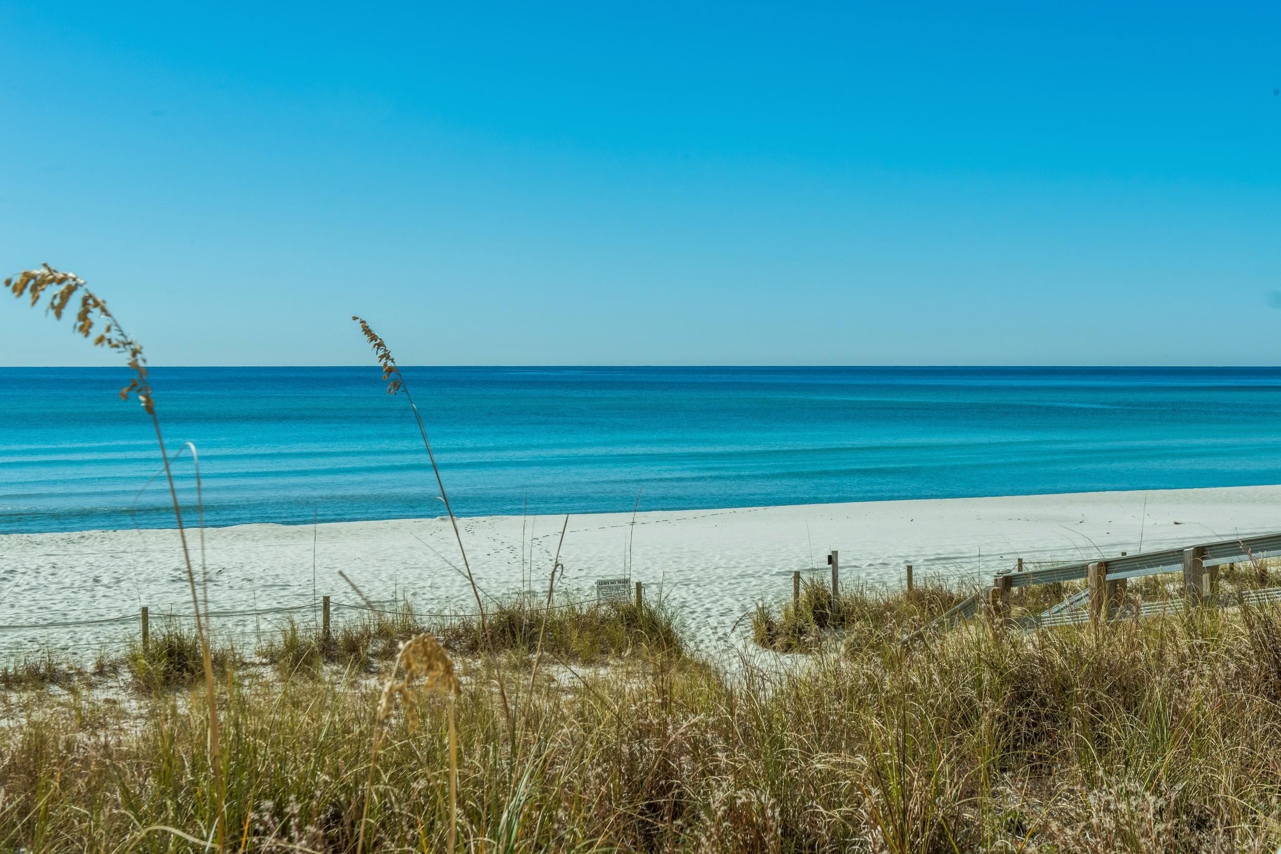 Ein Strand mit klarem, blauem Wasser, sandigem Ufer und etwas Gras im Vordergrund.