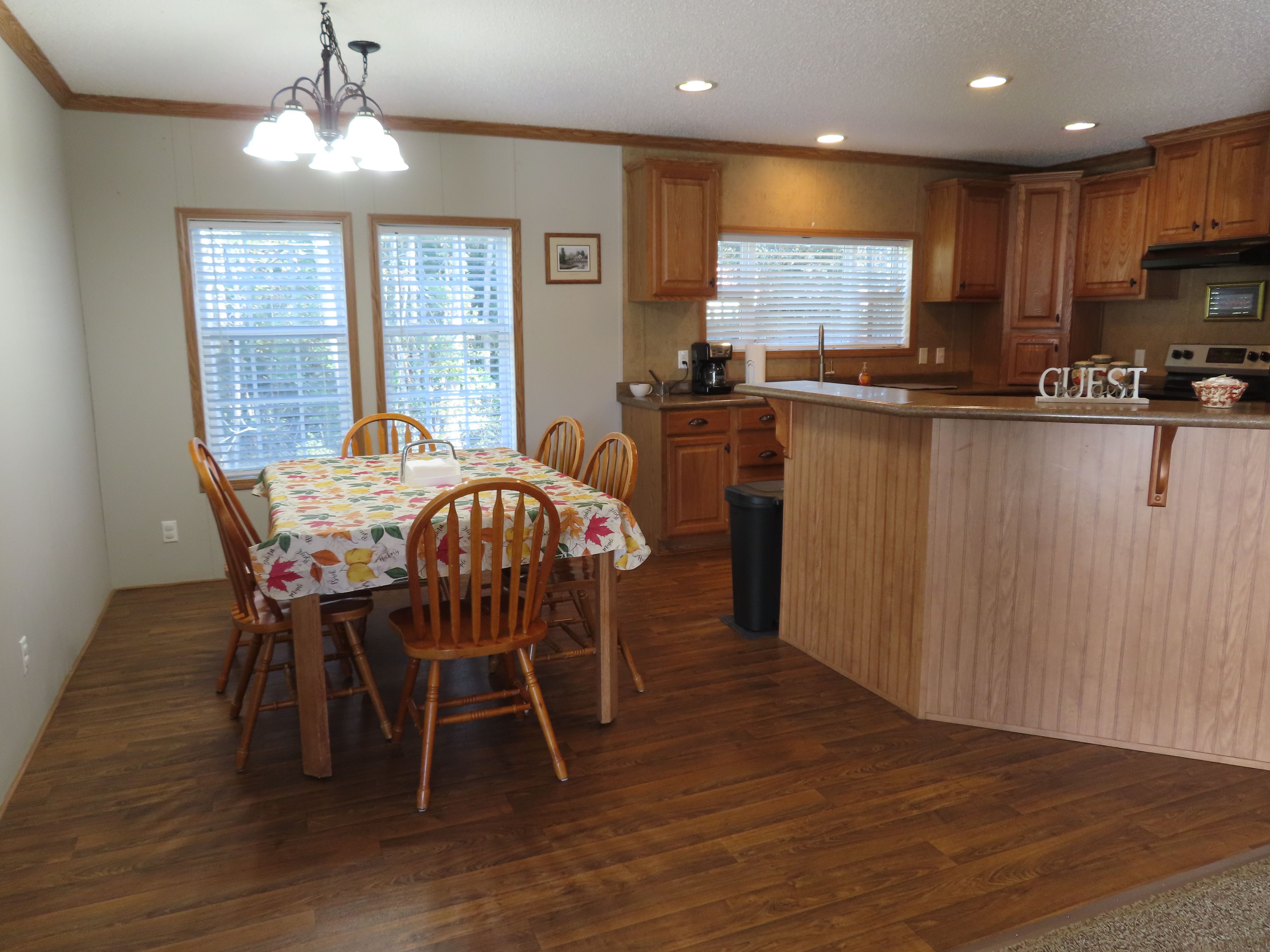 A kitchen with wooden cabinets, a dining table with chairs, a trash bin, and a sign that says 'GUEST'.