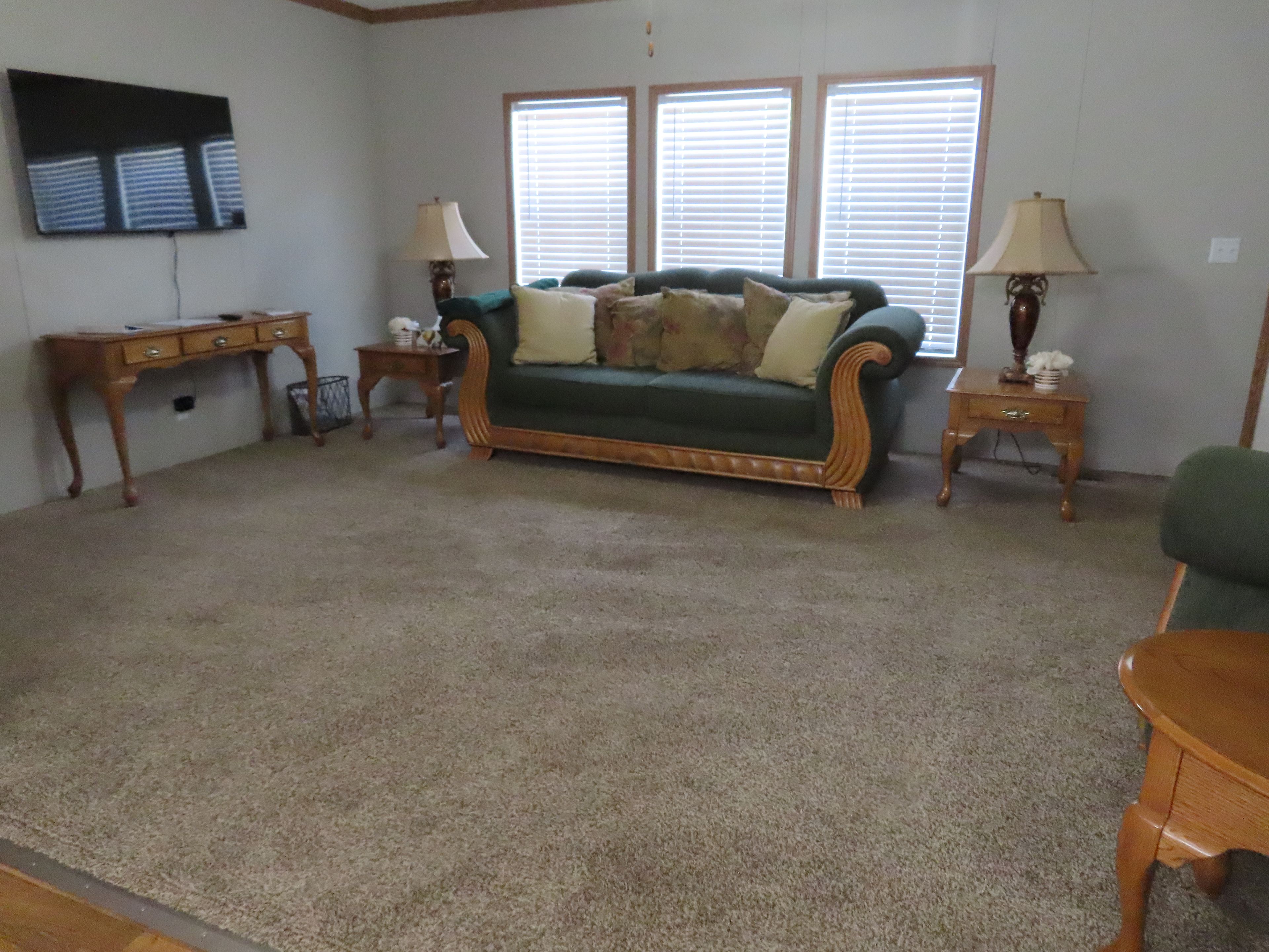 A living room with a green sofa, two side tables with lamps, a television on the wall, and a large window with blinds.