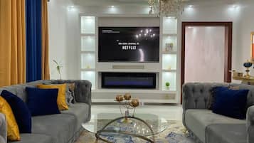 A modern living room with two grey sofas, a glass coffee table, and a flat-screen TV mounted on the wall.