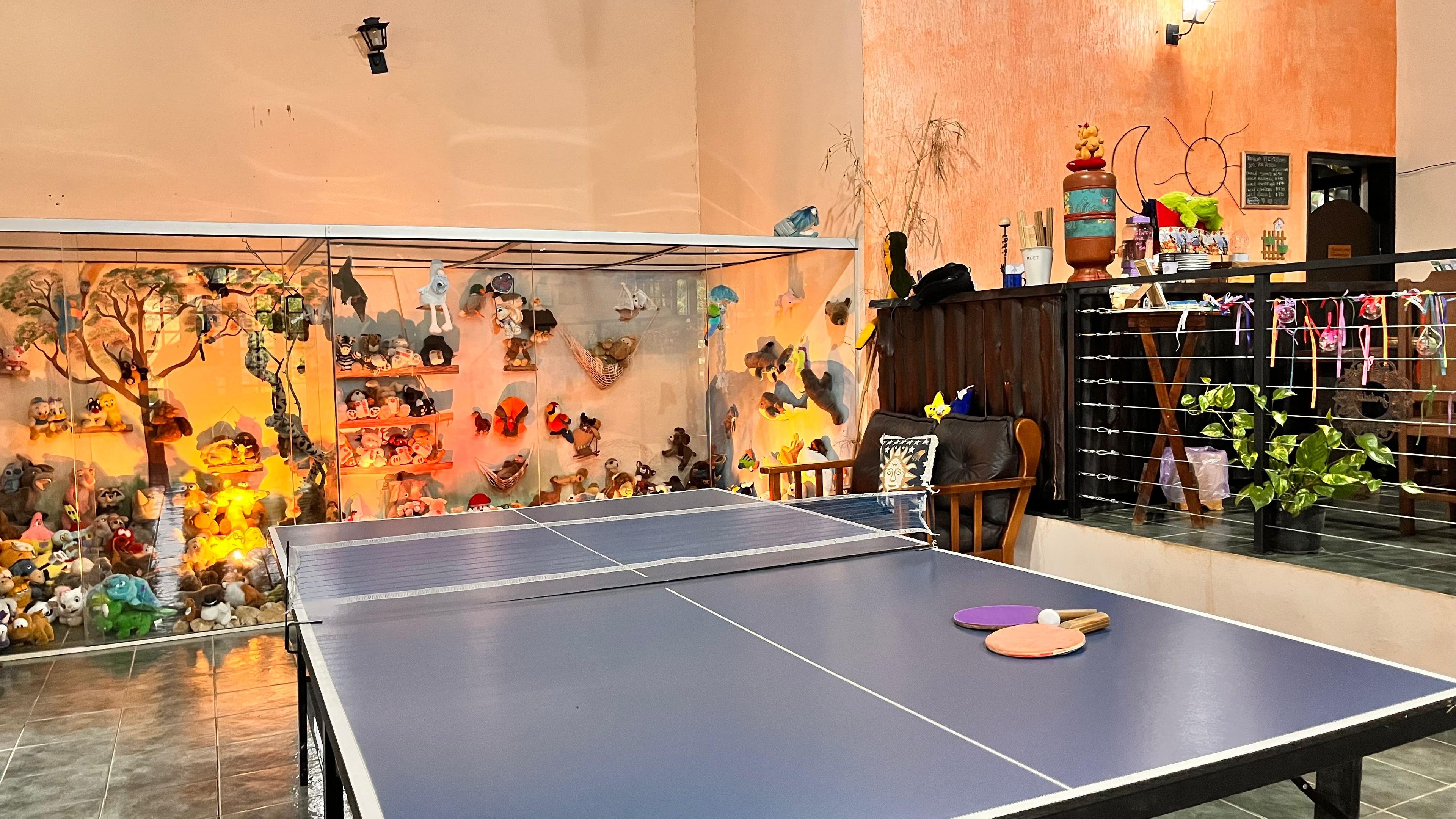 A ping pong table in a room with a wall decorated with various plush toys and a small seating area with a chair and a table.