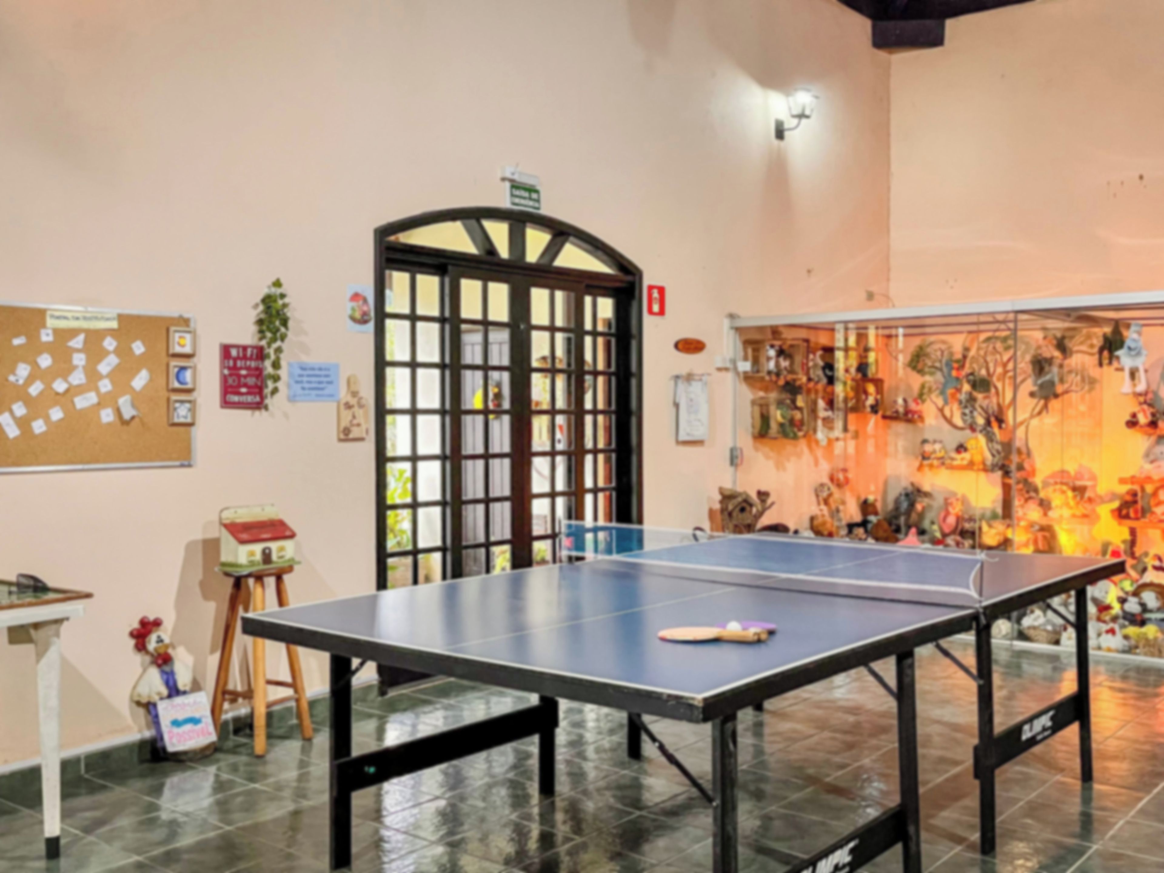 Mesa de pingue-pongue em um quarto com um painel de cortiça na parede, um banquinho pequeno e uma porta com janela.