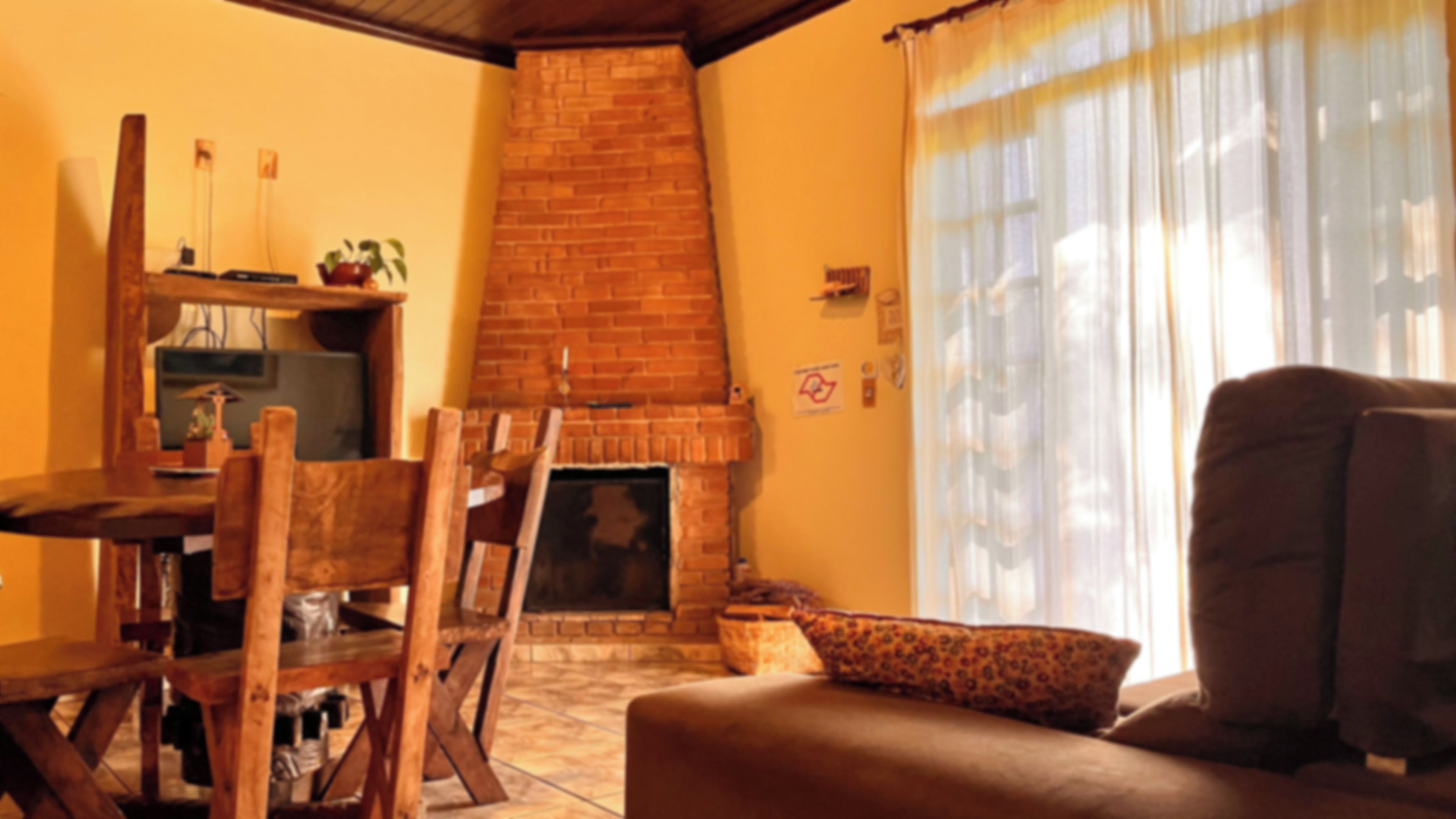 Ein Wohnzimmer mit Kamin aus Backstein, Holzmöbeln und einem großen Fenster mit transparenten Vorhängen.