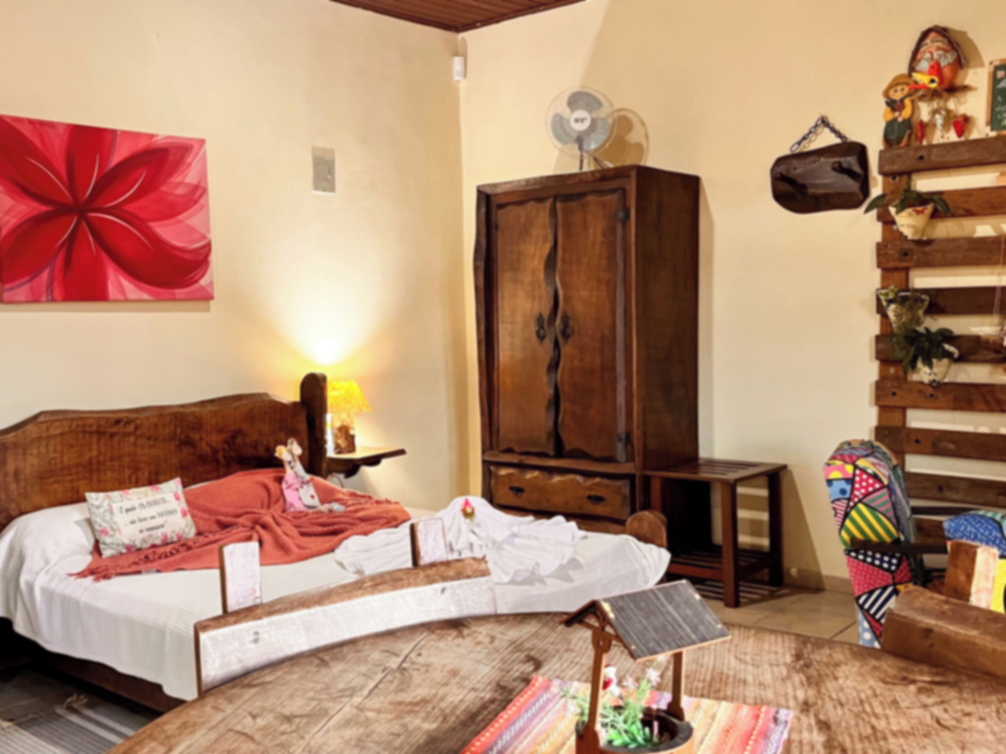 Quarto com cama, um armário de madeira, uma mesinha e uma cadeira colorida.