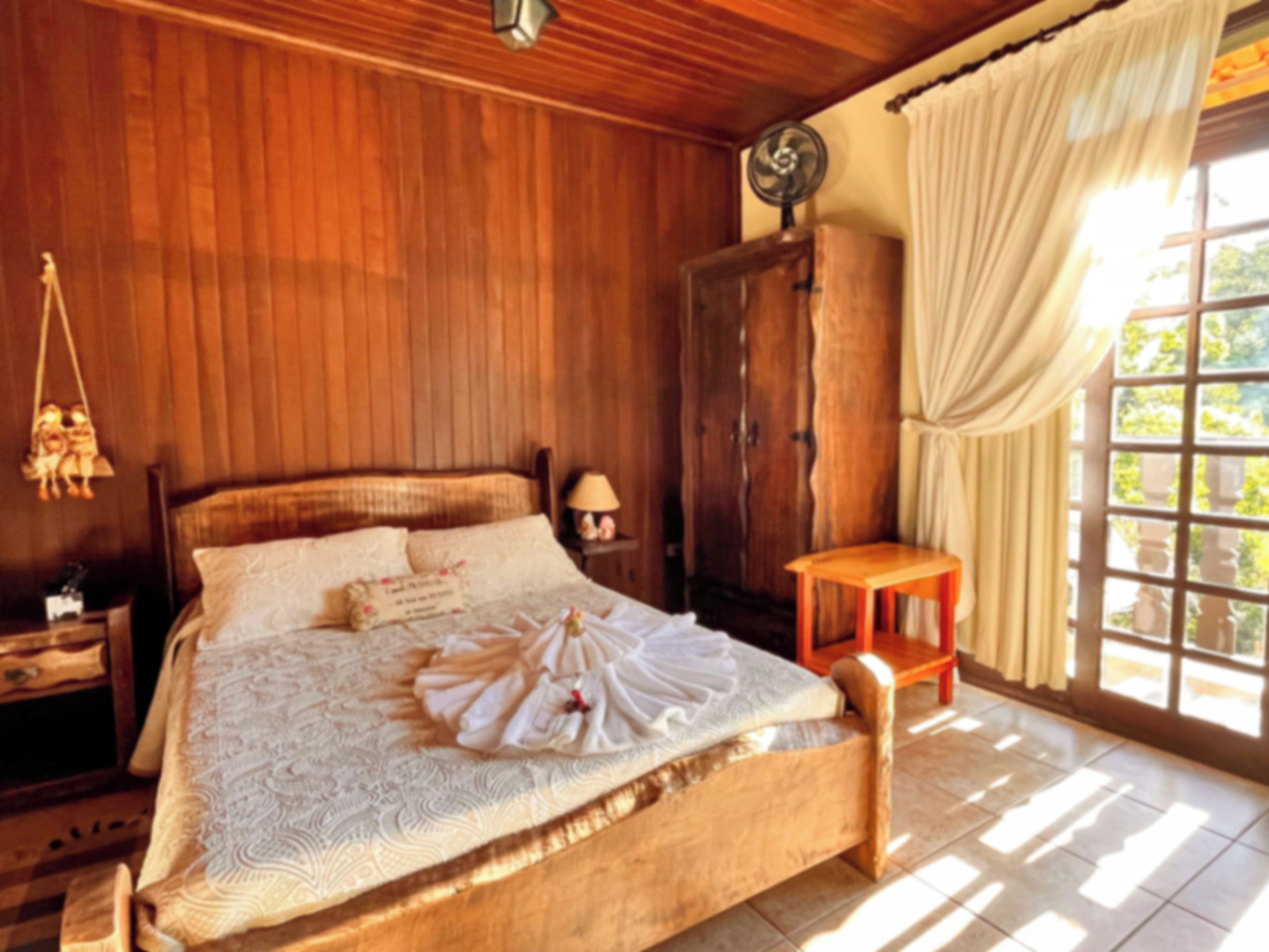 Uma cama com estrutura de madeira, com colcha e almofadas brancas, um criado-mudo com abajur, um guarda-roupa de madeira e uma janela com cortinas.