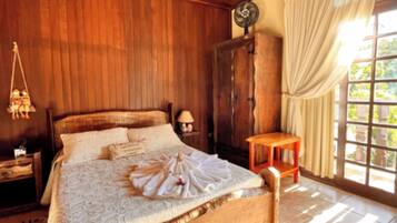 Uma cama com estrutura de madeira, com colcha e almofadas brancas, um criado-mudo com abajur, um guarda-roupa de madeira e uma janela com cortinas.