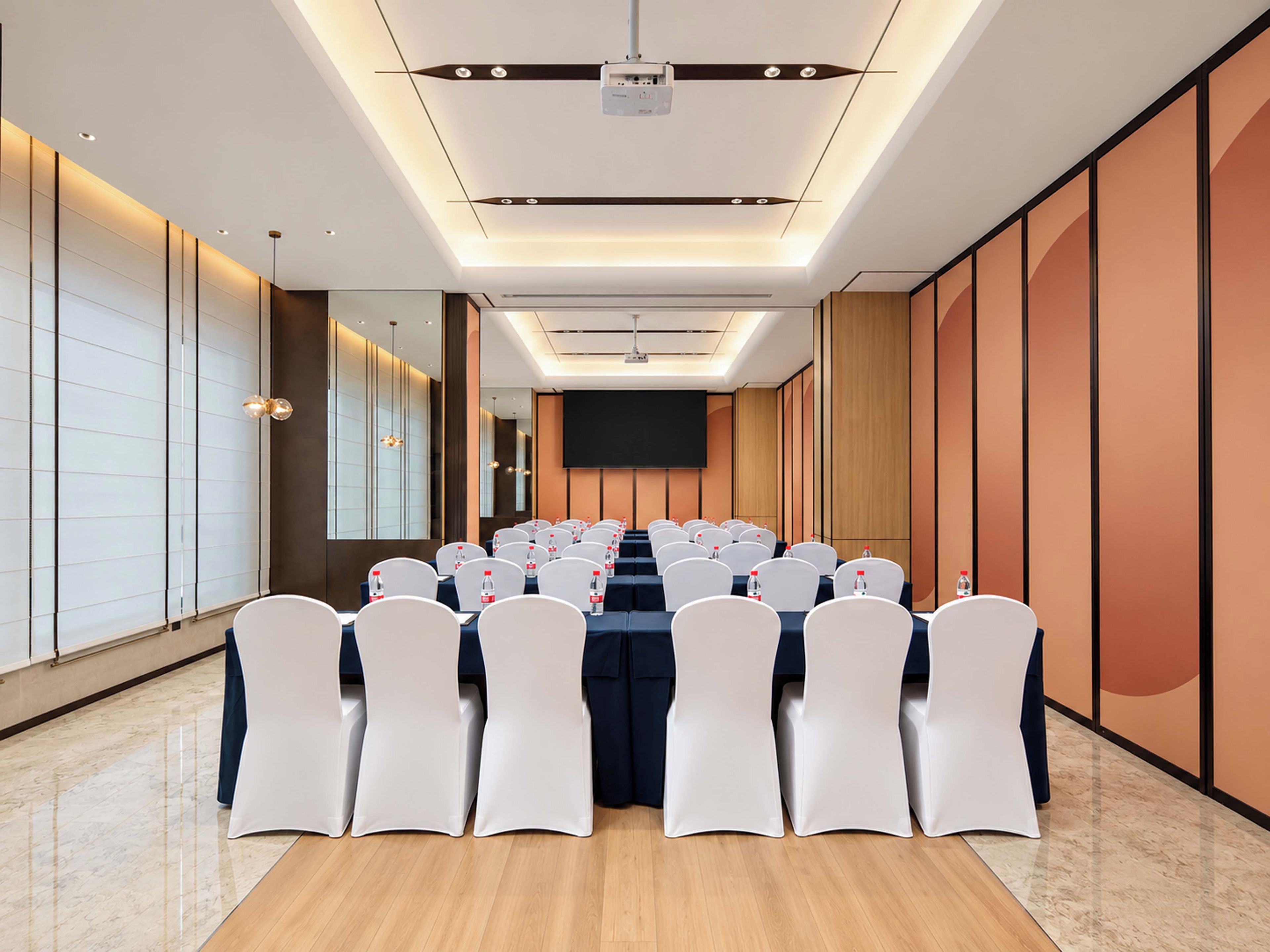 A conference room with rows of white chairs, a long table, and a projector screen at the front.