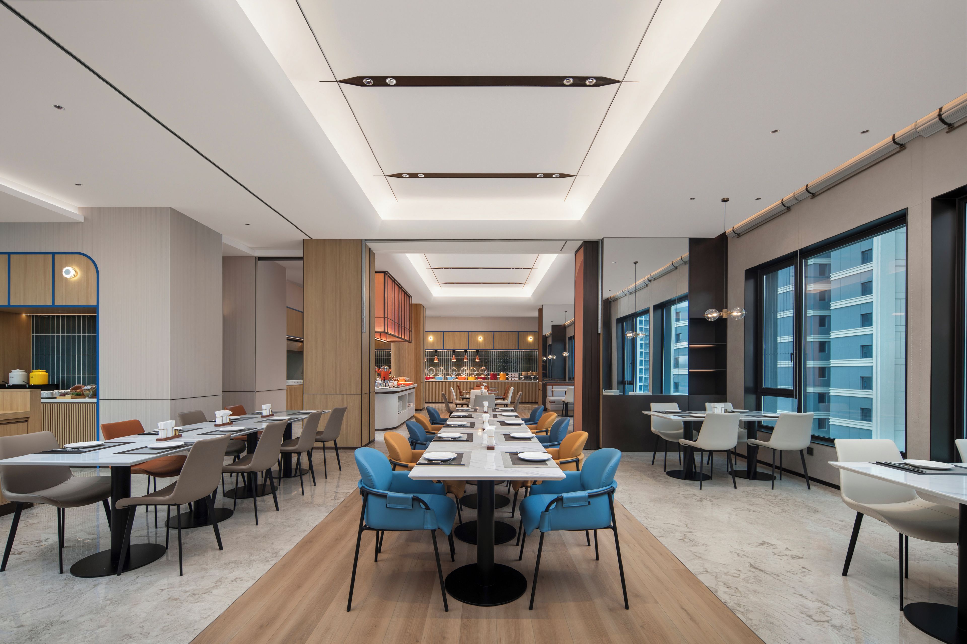 A modern dining area with long tables, chairs, and a kitchen in the background.